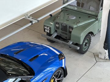 Een vintage groene Land Rover met kentekenplaat "HUE 166" staat naast een moderne blauwe sportwagen in een smetteloze garage en legt een scène vast die doet denken aan de reportage van het British Motor Museum.