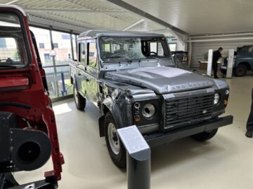 Een beschadigde Land Rover Defender met gebroken ruiten en een verbogen motorkap wordt tentoongesteld in een lichte tentoonstellingsruimte in het British Motor Museum. Het frame van een rood voertuig is links gedeeltelijk zichtbaar en ervoor staat een informatiebord.