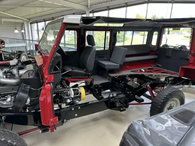 Gedeeltelijk geassembleerd rood terreinvoertuig met zichtbare interne componenten en interieur. Het voertuig, tentoongesteld in het British Motor Museum, wordt tentoongesteld in een goed verlichte showroom, terwijl andere voertuigen zichtbaar zijn door grote ramen.