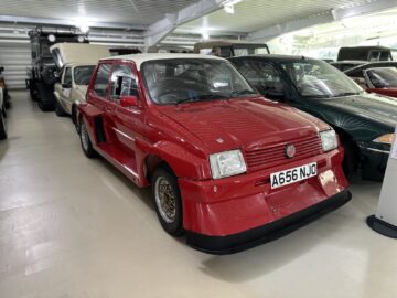 Een rood-witte hatchback met een 'A656 NJO'-kentekenplaat staat geparkeerd in een lichte, ruime showroom naast andere voertuigen, mogelijk klaargemaakt voor een reportage in het British Motor Museum.