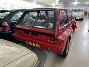 Een rode vintage auto staat binnen op een rij geparkeerd met andere voertuigen bij het British Motor Museum. Op het kenteken van de auto staat "A656 NJO", en op de achterruit zit een kleine sticker.