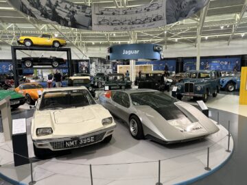 Een tentoonstelling van klassieke auto's in een tentoonstelling in het British Motor Museum, waaronder een witte sportwagen met het kenteken "NMT 335E" en een strakke zilveren conceptauto, met een Jaguar-bord zichtbaar op de achtergrond. Perfect voor iedereen die een bezoek plant of geïnteresseerd is in een autoreportage.