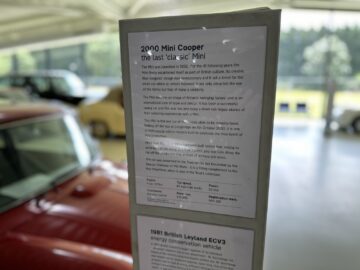 Een close-up van een informatiebord in het British Motor Museum beschrijft een Mini Cooper uit 2000, ook wel de laatste "klassieke" Mini genoemd. Auto's in de buurt en een onscherpe achtergrond voegen context toe aan deze reportage.