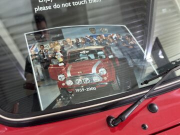 Foto van een rode auto in het British Motor Museum, met een gedrukte afbeelding onder de voorruit waarop een groep feestvierende mensen te zien is. Op de print is de tekst "1959-2000" zichtbaar, waarmee een nostalgisch moment uit de autogeschiedenis wordt vastgelegd.