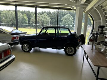 Een compacte, vintage zwarte auto die binnen geparkeerd staat bij grote ramen en met groen aan de buitenkant springt in het oog. Omhuld door andere klassieke auto's en een motormodel op een tafel, voelt het als een scène uit een reportage van het British Motor Museum die wacht op uw bezoek.