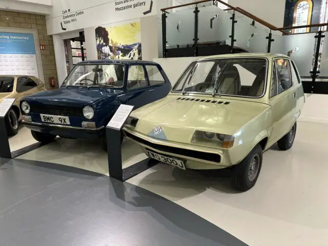 Op de binnententoonstelling van het British Motor Museum worden twee compacte oldtimers naast elkaar getoond in een reportage. De blauwe auto, met het opschrift 'BMC 9X', en de beige auto, met de aanduiding 'Bond', hebben elk informatieve plaquettes aan de voorkant die bezoekers kunnen bekijken.