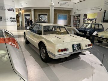 Een vintage witte auto met kenteken NMT 395E wordt tentoongesteld in een reportagesetting in het British Motor Museum, genesteld tussen andere klassieke voertuigen, waardoor het een must-see bezoek is voor liefhebbers.