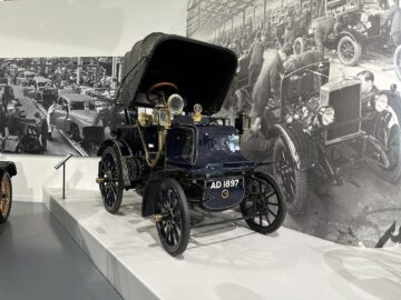 Een oldtimer met kenteken AD1897 staat tentoongesteld op een platform in het British Motor Museum. Een zwart-witte muurschildering van een oude autoproductiescène vormt een nostalgische achtergrond, perfect voor uw volgende bezoek of RAPPORTAGE.