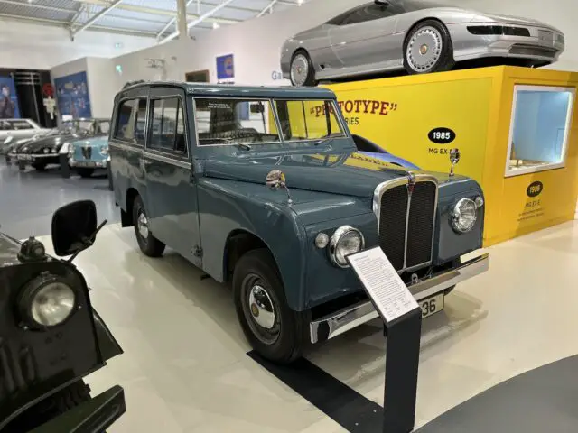 In het British Motor Museum wordt een blauwe vintage auto tentoongesteld in een museumsetting, geparkeerd naast een gele tentoonstellingsstand met een prototype automodel uit 1985. Andere auto's zijn zichtbaar op de achtergrond, wat de bezoekerservaring vergroot.