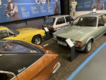 Een bezoek aan het British Motor Museum toont een tentoonstelling van oldtimers, waaronder een groene Ford, een witte Fiesta, een gele sportwagen en een bruin voertuig met een 'Wolseley'-badge. De geblokte vloer en informatieborden dragen bij aan de charme van elk bezoek.