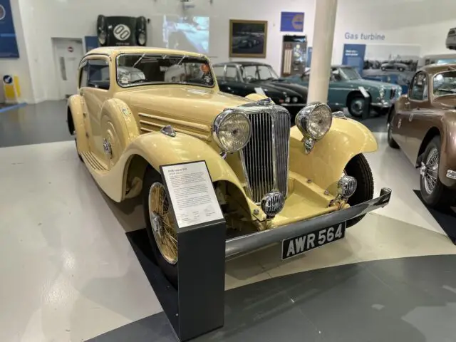 Een vintage gele auto met een Brits kenteken "AWR 564" wordt tentoongesteld in het British Motor Museum, omringd door andere klassieke auto's en informatieve displays. Als u een bezoek plant, kunt u een indrukwekkende reportage over de autogeschiedenis verwachten.