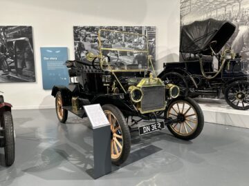 Een vintage zwarte auto met koperen armaturen en houten spaakwielen wordt tentoongesteld in het British Motor Museum. Op de achtergrond zijn informatieve plaquettes en historische foto's te zien, die diepte toevoegen aan deze tentoonstelling in reportagestijl die bij elk bezoek informatief zal zijn.