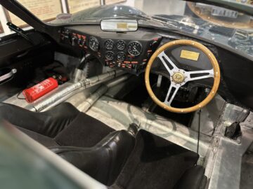 Interieur van een vintage raceauto in het British Motor Museum, met een stuur met houten rand, een dashboard met meerdere meters, twee zwarte kuipstoelen en een rode brandblusser op de vloer. Bezoek deze tentoonstelling voor een exclusieve reportage-ervaring.