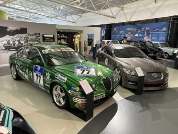 Twee Jaguar-raceauto's tentoongesteld in het British Motor Museum, één groene en één grijze, elk met sponsorstickers en het nummer 77. Op de achtergrond staan historische race-exposities en memorabilia, die een rijke bezoekervaring bieden voor elke autosportliefhebber.