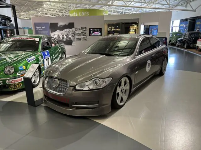 Een slanke, zilveren sportwagen wordt tentoongesteld in een overdekte autotentoonstelling in het British Motor Museum, omringd door andere vintage- en racevoertuigen. Bezoek en verdiep je in deze prachtige collectie.