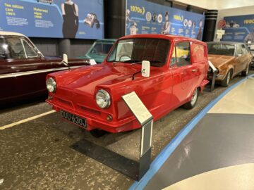 Een rode vintage driewielige bestelwagen, met het kenteken "DUU 630J", wordt tentoongesteld in het British Motor Museum. Een informatiebord begeleidt dit klassieke voertuig en biedt bezoekers een kijkje in de geschiedenis ervan.