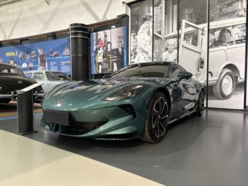 In een showroom wordt een strakke, moderne groene sportwagen tentoongesteld met historische autoposters op de achtergrond, die doen denken aan een reportage uit het British Motor Museum.