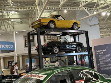 Een gele cabriolet wordt tentoongesteld boven een zwarte auto op een metalen rek in het British Motor Museum. Op de achtergrond zijn andere voertuigen en tentoonstellingsstukken zichtbaar, waaronder een groene sportwagen op de voorgrond.