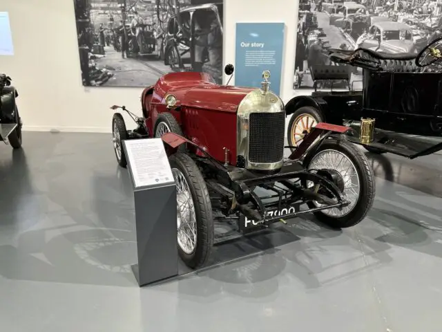 Een vintage rode raceauto met een grote grille en spaakwielen wordt tentoongesteld in het British Motor Museum. Voor de auto staat een beschrijvende plaquette, terwijl op de achtergrond historische autoafbeeldingen de muur sieren en bezoekers een boeiend kijkje in de autogeschiedenis bieden.