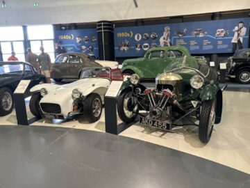 Een bezoek aan het British Motor Museum onthult een voortreffelijke tentoonstelling van antieke auto's uit de jaren vijftig, waaronder een witte sportwagen met het opschrift NRM 7 en een groen driewielig voertuig met kenteken AKD 455. Gedetailleerde informatie wordt gegeven op panelen, waardoor dit diepte krijgt. boeiende autoreportage.