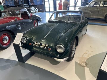 Een groene klassieke sportwagen met kenteken "287 JRT" wordt tentoongesteld in een showroom in het British Motor Museum, omringd door andere vintage voertuigen. Naast de auto staan informatieborden met informatie voor bezoekers.