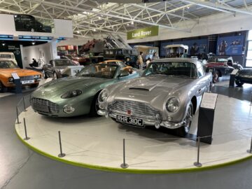 Twee klassieke auto's, een groene coupé en een zilveren cabriolet, staan tentoongesteld op een verhoogd platform in het British Motor Museum. Op de achtergrond is een Land Rover-sectie zichtbaar. Deze tentoonstelling is perfect voor iedereen die van plan is een bezoek te brengen aan of een reportage te schrijven over de autogeschiedenis.