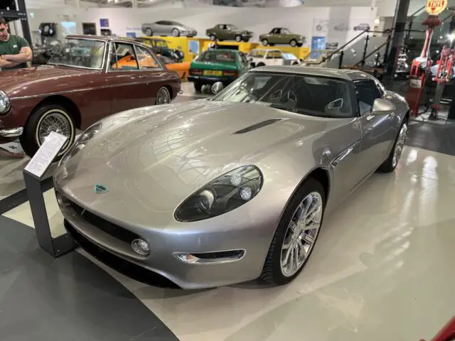 Een zilveren sportwagen wordt tentoongesteld in een showroom naast andere voertuigen in het British Motor Museum. Ernaast staat een bord met informatie over de auto, dat uitnodigt tot een bezoek voor meer informatie.