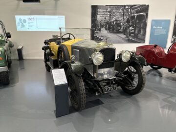 Een oldtimer met cabriokap wordt tentoongesteld in de tentoonstelling van het British Motor Museum. De auto heeft een geel-zwarte carrosserie met historische details en wordt vergezeld door een informatief plaatje.