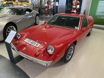 Een vintage rode Lotus-sportwagen met kenteken "AR0401K" wordt binnen tentoongesteld in het British Motor Museum, naast een informatiebord met details over de geschiedenis ervan.