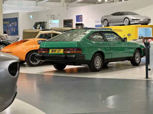 Een groene vintage sportwagen met een "BHP 2T" kenteken wordt tentoongesteld in het British Motor Museum, samen met een oranje auto op de achtergrond en diverse andere auto's. Als u er langskomt, heeft u het gevoel dat u in een boeiende reportage over de autogeschiedenis stapt.