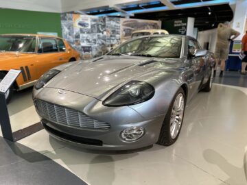 Een zilveren sportwagen wordt tentoongesteld in een museumomgeving naast een oranje auto, met een tentoonstellingsbord vlakbij en verschillende oldtimerfoto's aan de muur op de achtergrond, die de essentie van een bezoek aan het British Motor Museum weergeven.