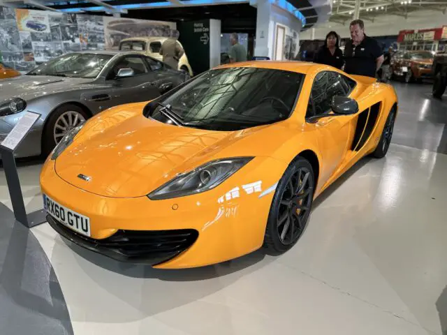 Een feloranje sportwagen met kenteken "RX60 GTU" is samen met andere voertuigen te zien in een showroom in het British Motor Museum.