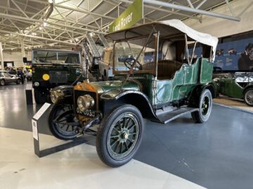 Een vintage groene auto met een wit dak tentoongesteld in het British Motor Museum. Een uithangbord en andere vintage voertuigen bevinden zich op de achtergrond en geven een gevoel van tijdloze elegantie en geschiedenis weer.