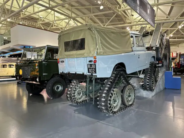 Een groen 4x4-voertuig staat geparkeerd naast een lichtblauwe 4x4 met sporen op de achterwielen, die binnen onder industriële verlichting in het British Motor Museum wordt tentoongesteld.