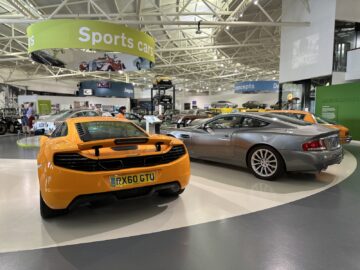 Een museumtentoonstelling in het British Motor Museum omvat een feloranje sportwagen en een slanke zilveren auto, tentoongesteld tussen andere voertuigen onder de vlag 'Sportwagens', wat een spannend bezoek biedt voor autoliefhebbers.