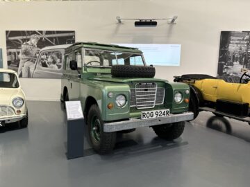 Een groene Land Rover met kenteken "R0G 924R" staat binnen geparkeerd in het British Motor Museum, tussen een gele auto en een crèmekleurige auto. Aan de muur achter de voertuigen hangen twee zwart-witfoto's, die de historische reportage van het museum versterken.