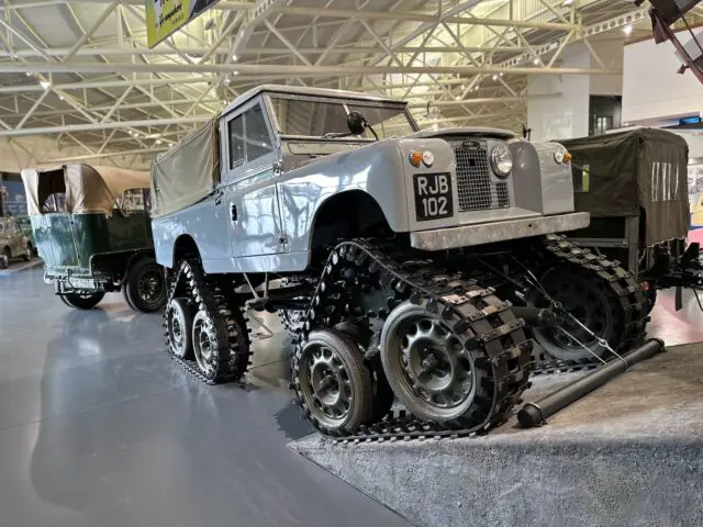 Een terreinwagen uitgerust met doorlopende spoorombouw staat tentoongesteld in een tentoonstellingshal met hoge plafonds en andere militaire voertuigen op de achtergrond, een scène die doet denken aan gedetailleerde reportages in het British Motor Museum.