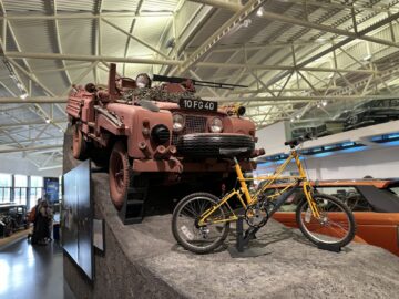 Een rood terreinvoertuig met het kenteken "10 FG 40" wordt tentoongesteld op een rotsachtige helling in het British Motor Museum, achter een gele fiets. Het plafond is voorzien van zichtbare balken en verlichting, wat bijdraagt aan de authentieke sfeer van dit intrigerende bezoek.