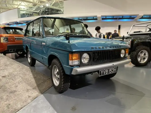 Een vintage blauwe Range Rover met kenteken YVB 153H wordt binnenshuis tentoongesteld in het British Motor Museum, omringd door andere voertuigen en mensen op de achtergrond.