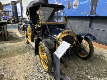 Een vintage blauw-zwarte auto met gele wielen wordt binnenshuis tentoongesteld in het British Motor Museum. De auto heeft een schuine voorruit en informatieborden ervoor. Op de achtergrond maakt een blauwe muur met verschillende exposities het tafereel compleet, terwijl bezoekers hun bezoek door de galerij voortzetten.