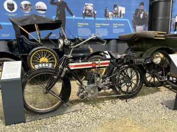 Een vintage motorfiets tentoongesteld in een museumtentoonstelling in het British Motor Museum, met andere klassieke voertuigen en informatieborden op de achtergrond, wacht op bezoek om hun eigen reportagewaardige momenten vast te leggen.