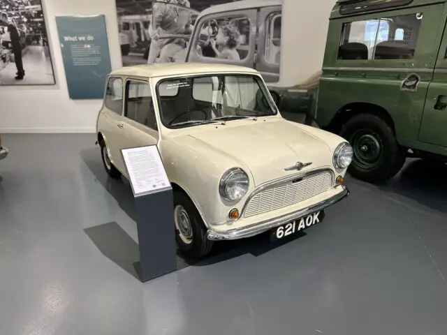 In het British Motor Museum wordt binnen een witte klassieke auto met kenteken "621 AOK" tentoongesteld naast een groen voertuig en een informatief plaatje. Machine- en historische foto's zijn zichtbaar op de achtergrond en leggen de essentie van de autogeschiedenis vast in deze gedetailleerde reportage.