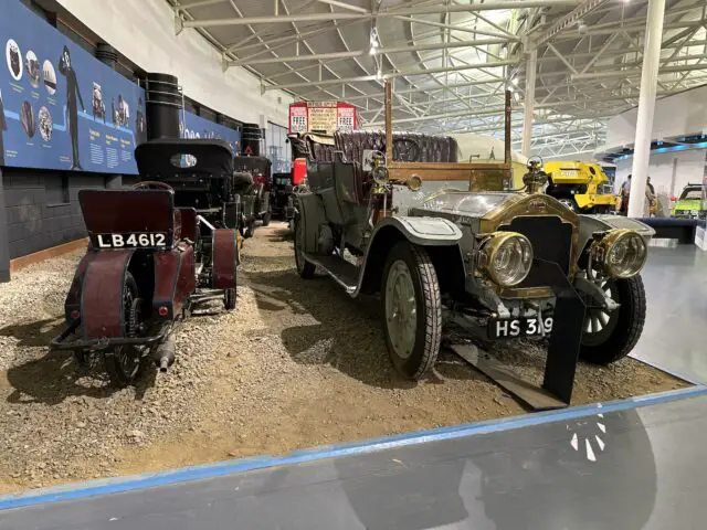 Vintage voertuigen, waaronder een oude auto met kenteken HS 319 en een motorfiets met kenteken LB 4612, worden binnenshuis tentoongesteld op een grindoppervlak in het British Motor Museum.