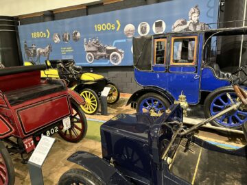 In het British Motor Museum zijn oldtimers uit de jaren 1890 tot 1900 te zien, met een rode auto, een gele auto en een blauwe auto, met informatieve panelen op de achtergrond die gedetailleerde reportages over hun geschiedenis bieden.