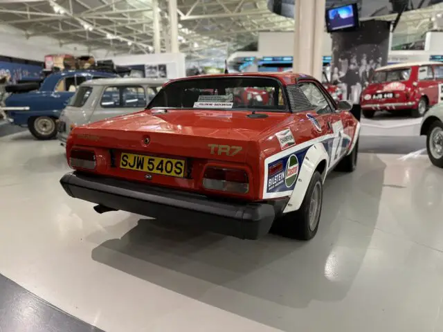 Een klassieke rode Triumph TR7-rallyauto met kenteken SJW 540S wordt tentoongesteld in het British Motor Museum, omringd door andere vintage voertuigen en auto-memorabilia. Deze voortreffelijke tentoonstelling is een must-see voor elk bezoek en neemt je mee terug in de tijd via een prachtige reportage over de autogeschiedenis.