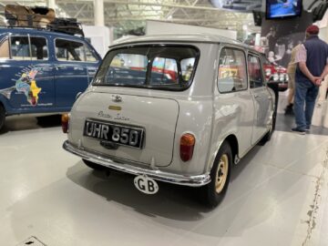 Een achteraanzicht van een lichtgrijze Austin Mini-auto met kenteken UHR 850, tentoongesteld in een binnententoonstelling in het British Motor Museum. Vlakbij is nog een blauwe auto zichtbaar, en mensen observeren de voertuigen in deze fascinerende reportage over de autogeschiedenis.