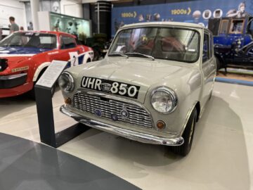 Een klassieke witte Mini Cooper met kenteken "UHR 850" is te zien in het British Motor Museum, omringd door andere vintage voertuigen en informatieve tentoonstellingen. Tijdens mijn bezoek vond ik vooral de gedetailleerde reportage over deze iconische auto fascinerend.