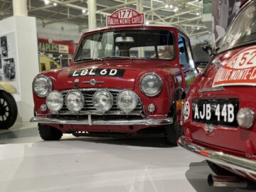 Twee rode klassieke Mini Coopers tentoongesteld, elk voorzien van kentekenplaten en rallyautostickers, waaronder 'Rallye Monte-Carlo'. De auto op de voorgrond heeft kenteken LBL 6D. Dit boeiende tafereel maakt deel uit van ons bezoek aan het British Motor Museum voor een spannende reportage over de autogeschiedenis.