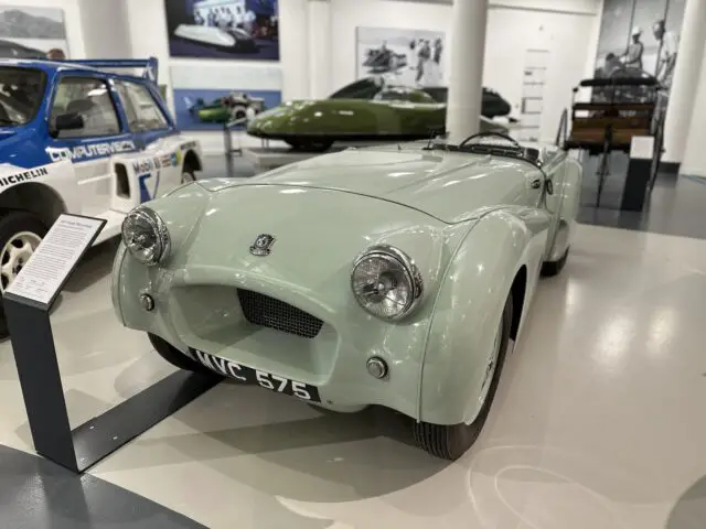 Een vintage lichtgroene auto met kentekenplaat "WC 575" wordt trots tentoongesteld in het British Motor Museum, omringd door andere voertuigen en informatieve borden. Deze fascinerende tentoonstelling is een must-see voor elk bezoek dat in de autogeschiedenis wil duiken.