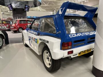 Een wit-blauwe rallyauto vertoont prominente merken, waaronder Computervision en Michelin, in een museumomgeving. De Metro 6R4, met zijn grote achterspoiler, maakt deel uit van de collectie van het British Motor Museum. Bezoek deze iconische tentoonstelling voor een boeiende autoreportage.